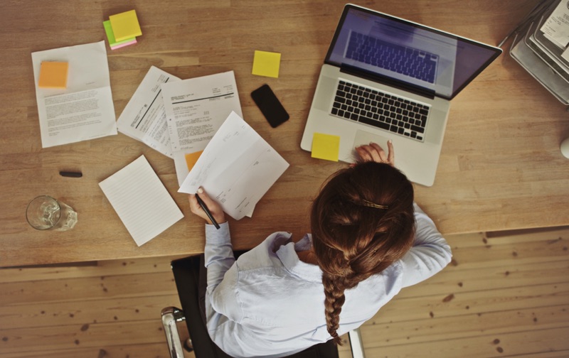 Mujer sentada en un escritorio de madera lleno de papeles, bloc de notas y una laptop