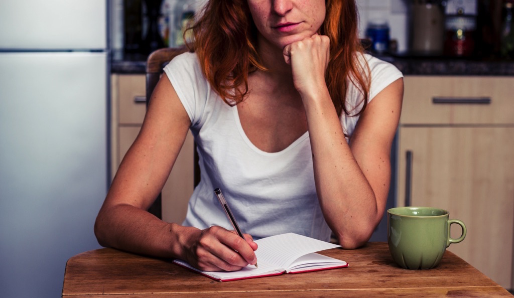 Mujer pelirroja haciendo un listado en una libreta con una pluma mientras toma una taza de café