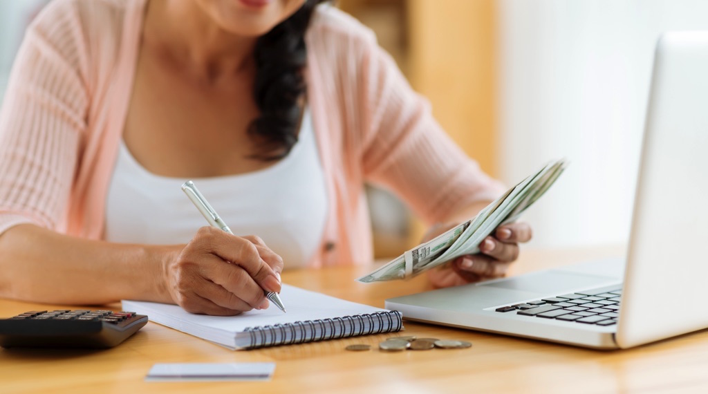 Mujer haciendo un presupuesto en una libreta con calculadora, dinero y laptop