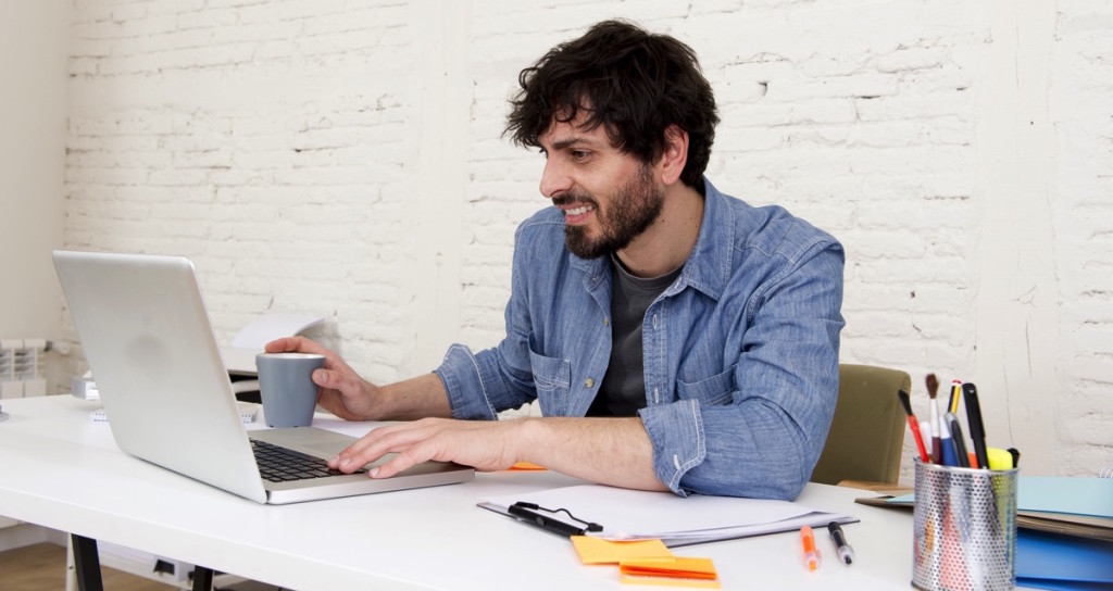 Persona adulta utilizando una laptop en su escritorio mientras toma una taza de café